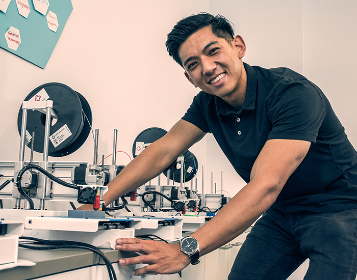 A student in a device engineering lab bending over his work and smiling. He has a black shirt and jeans on.